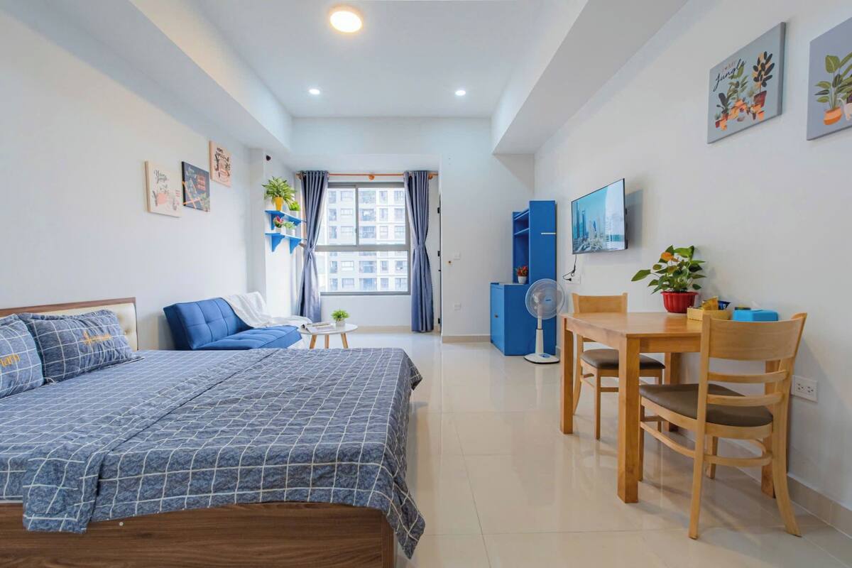 Ein modernes Schlafzimmer mit einem Bett, einem kleinen Tisch und einem Sessel. Ein Fenster mit Vorhängen, ein Fernsehgerät an der Wand und eine blaue Kommode sind ebenfalls vorhanden.