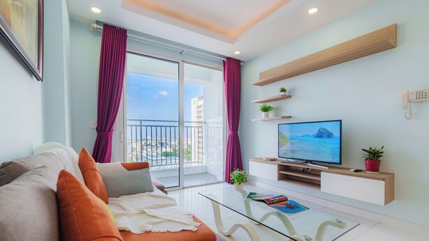 Una sala de estar moderna con un sofá, una mesa de centro de vidrio, un televisor de pantalla plana y un balcón con cortinas moradas.