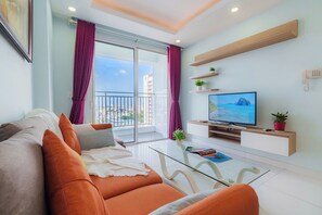 Ein modernes Wohnzimmer mit einer Couch, einem Glastisch, einem Flachbildfernseher und einem Balkon mit violetten Vorhängen.