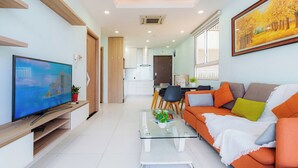 Una sala de estar moderna con televisor de pantalla plana, sofá naranja, mesa de centro de vidrio y una zona de comedor al fondo.