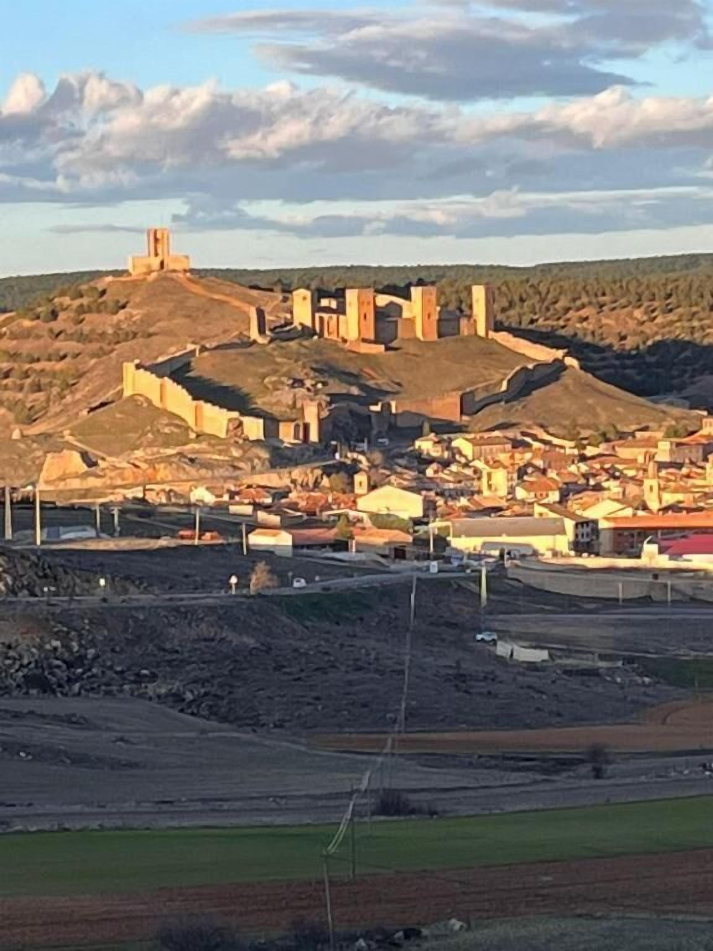 Un pueblo histórico con un castillo imponente en una colina, rodeado de viviendas y campos abiertos.