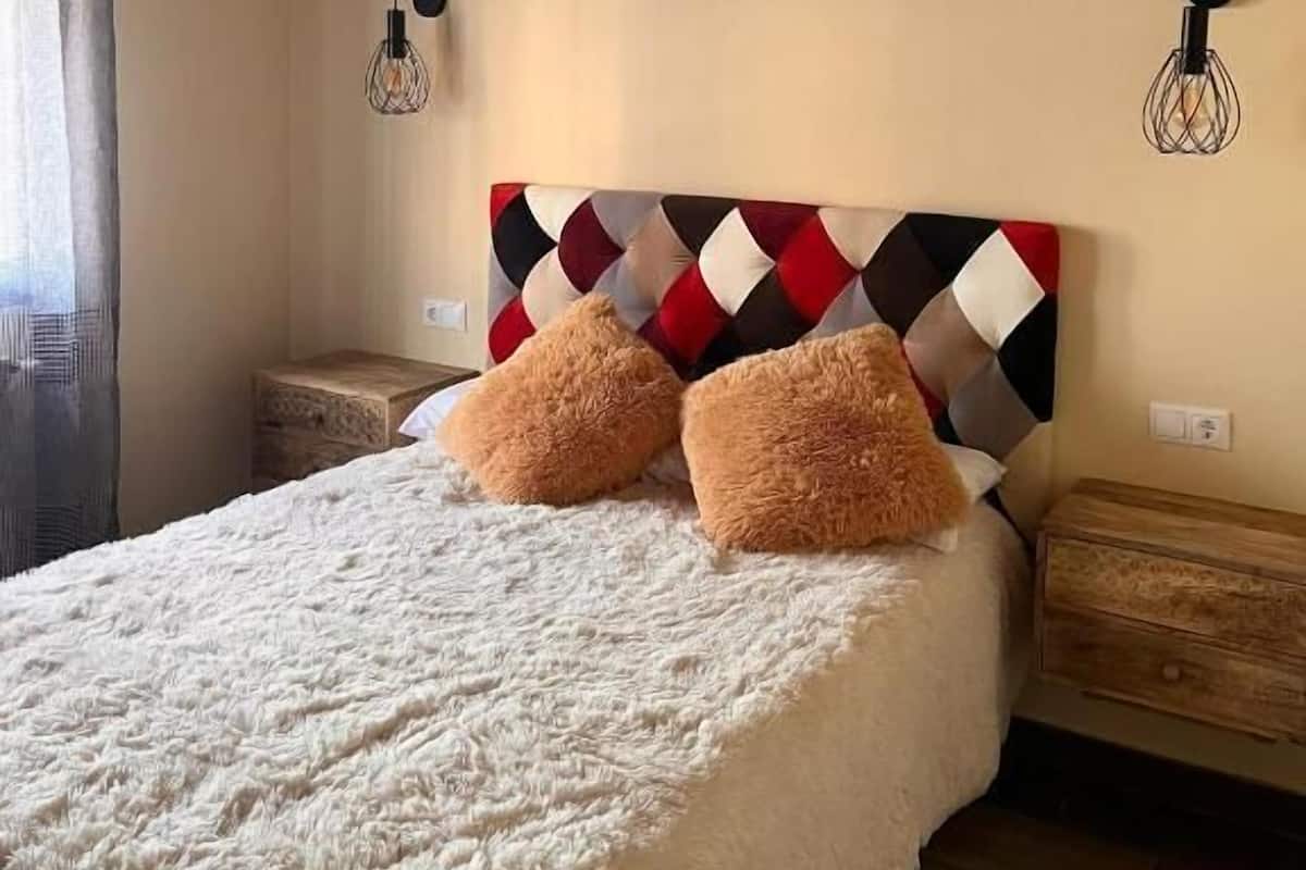 A bedroom with a bed, two fluffy pillows, a patterned headboard, bedside tables, and wall-mounted light fixtures.