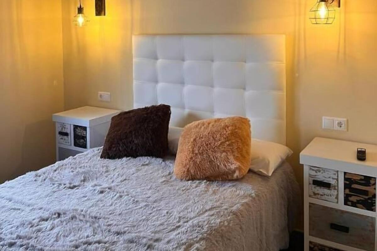 A bedroom with a tufted headboard, two bedside tables, a fluffy white bedspread, and two wall-mounted light fixtures.