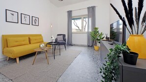A modern living room with a yellow sofa, a grey rug, a small round table, a grey armchair, a TV, and a window with grey curtains.