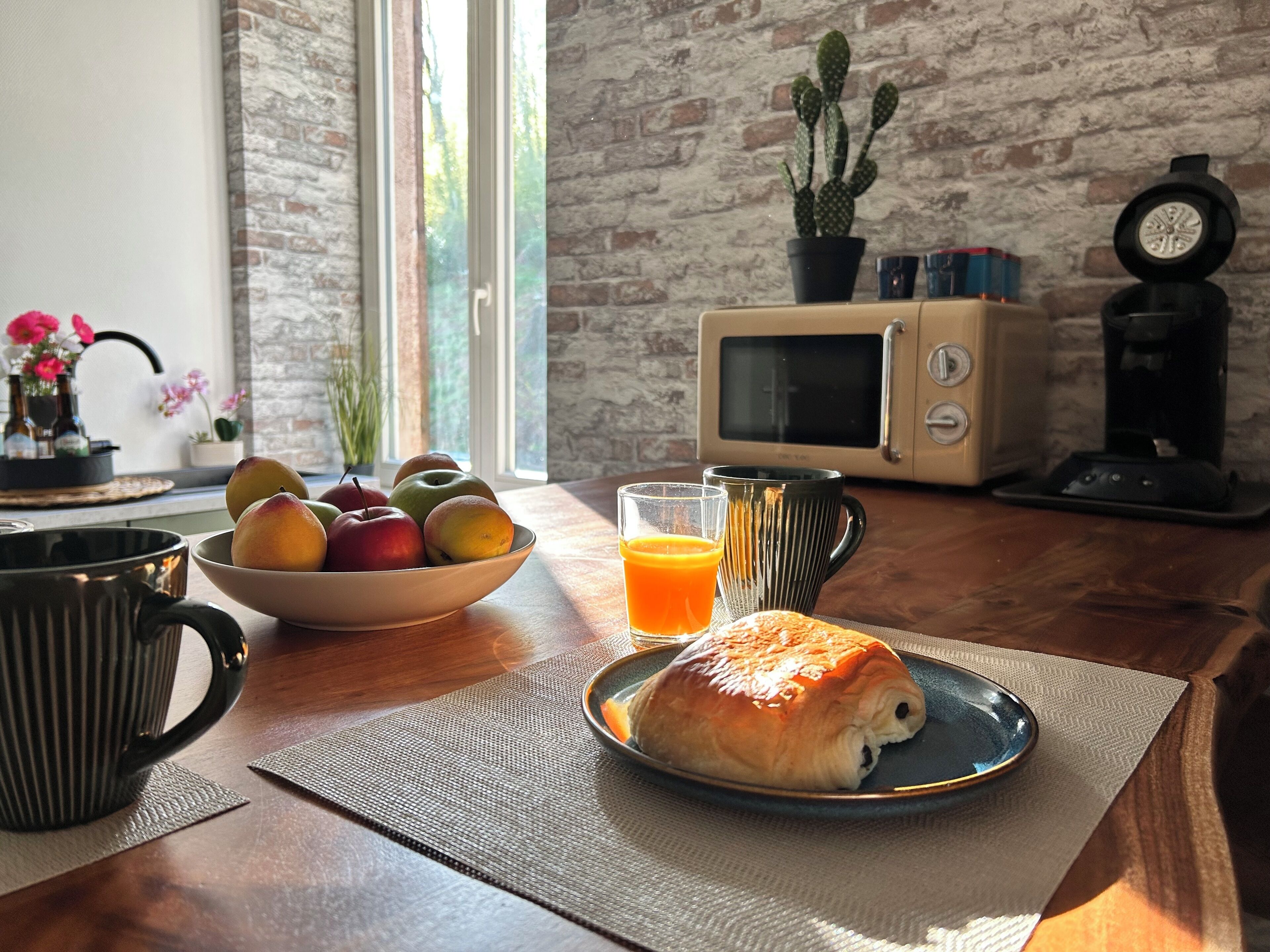 Une table en bois dressée pour le petit-déjeuner, avec un bol de fruits, un verre de jus d’orange, un croissant et un four à micro-ondes.