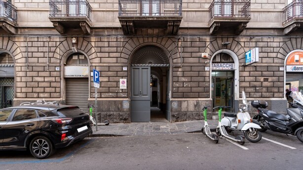 A street view with a parked car, scooters, and a building with a closed shop. - GuestHost - Central apartment Caronda Compact (Catania)