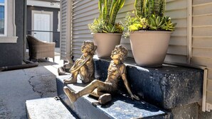 Deux statues de bronze représentant des enfants assis sur une corniche en béton, avec des plantes en pot en arrière-plan.