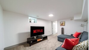 Ein Wohnzimmer mit einem Flachbildfernseher auf einem Schrank, einem grauen Sofa mit roten Kissen und einem kleinen Fenster mit einem Bilderrahmen.