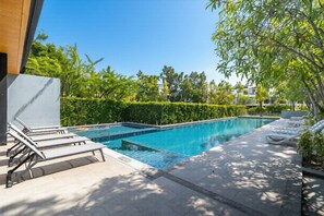 A rectangular swimming pool with a tiled deck, surrounded by greenery and a clear blue sky.