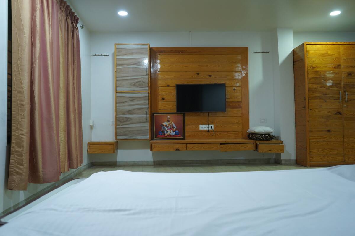 A bedroom with a bed, a wooden wardrobe, a television mounted on the wall, and a small cabinet below the TV.