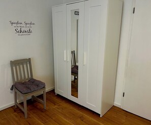 Une pièce avec un plancher en bois, une chaise dotée d’un coussin à motifs et une armoire blanche avec un miroir.