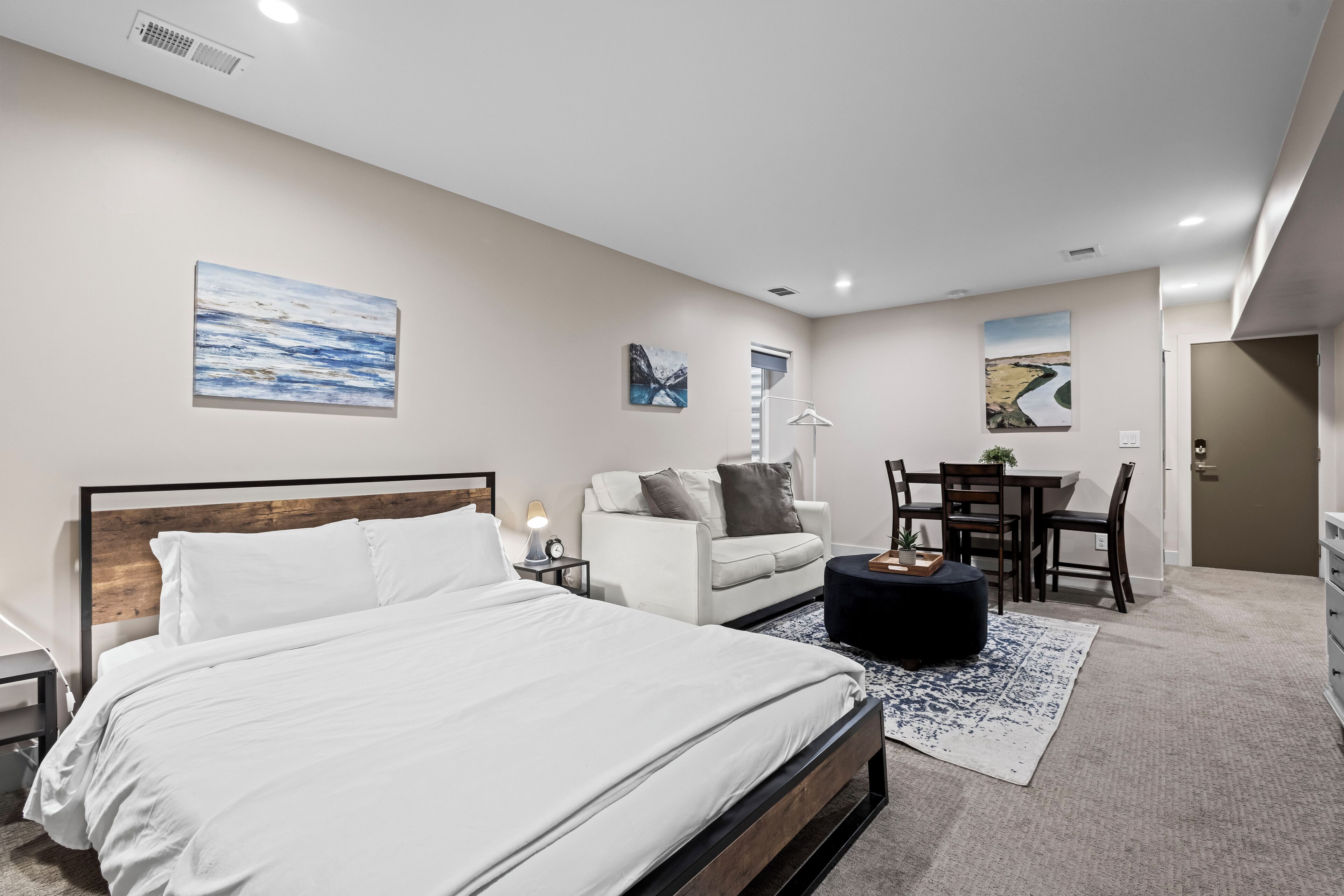 A modern bedroom with a large bed, a white sofa, a dining table, and artwork on the walls.