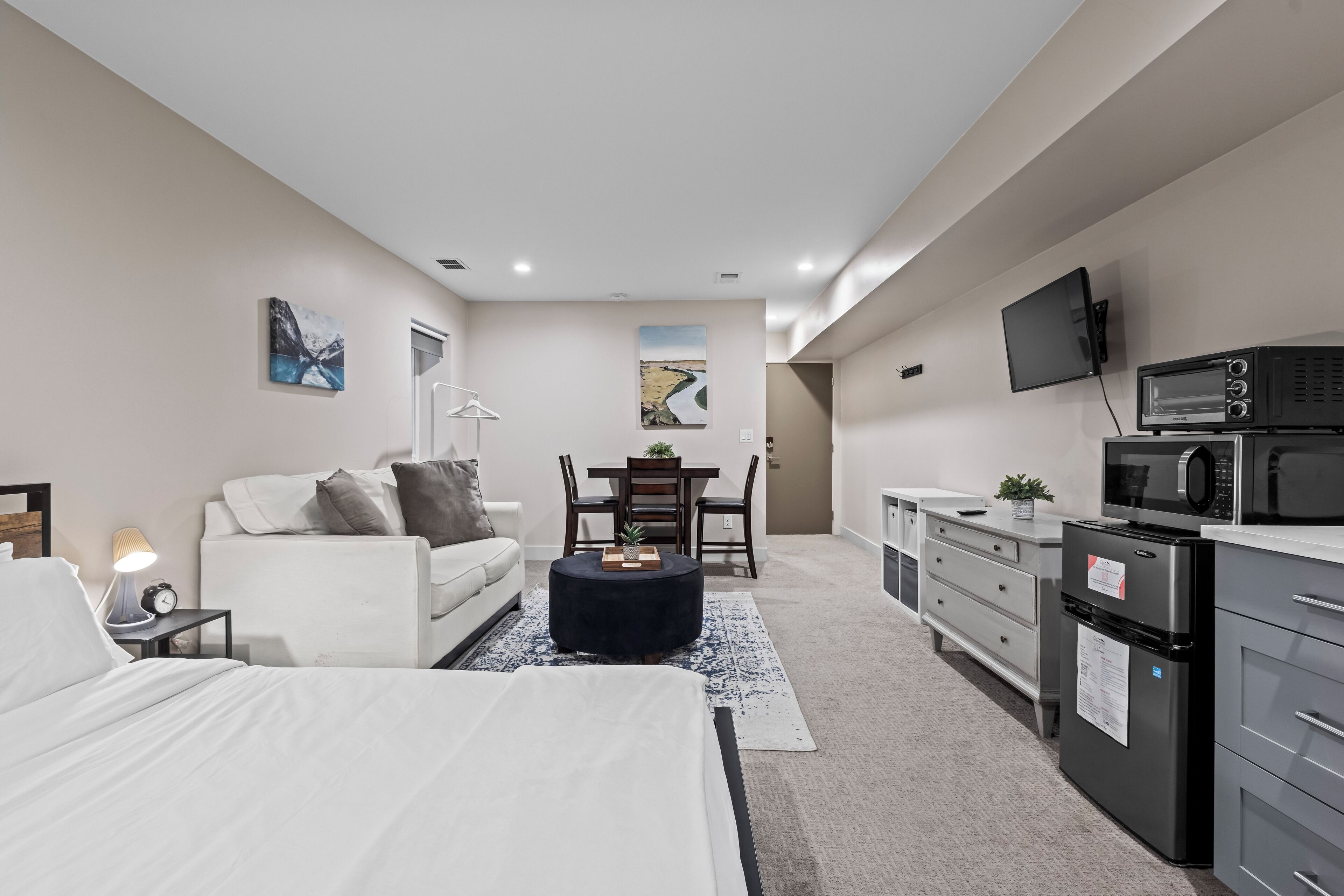 A modern, well-lit room with a bed, a white sofa, a dining table with chairs, a small kitchen area with a microwave and refrigerator, and artwork on the walls.