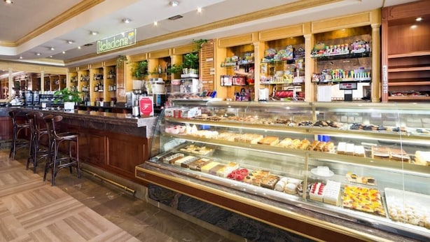 Una panadería con una variedad de pasteles y postres exhibidos en una vitrina, una barra de café con un letrero que dice "Café", y un área de estar con taburetes.