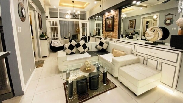 Una sala de estar moderna con un sofá blanco, una mesa de centro de vidrio y una zona de cocina con encimera blanca y un espejo grande.