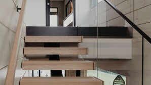 Un escalier moderne avec des marches en bois et une rampe en verre, menant à un espace situé au deuxième étage et offrant une vue sur l’extérieur.