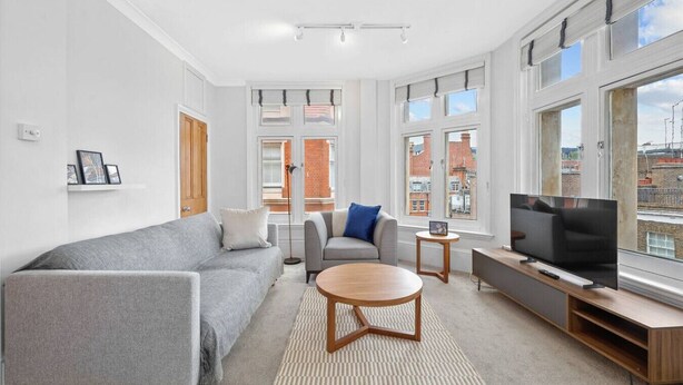 A modern living room with a grey sofa, a smaller grey armchair, a round wooden coffee table, a TV on a wooden stand, and large windows with a city view.