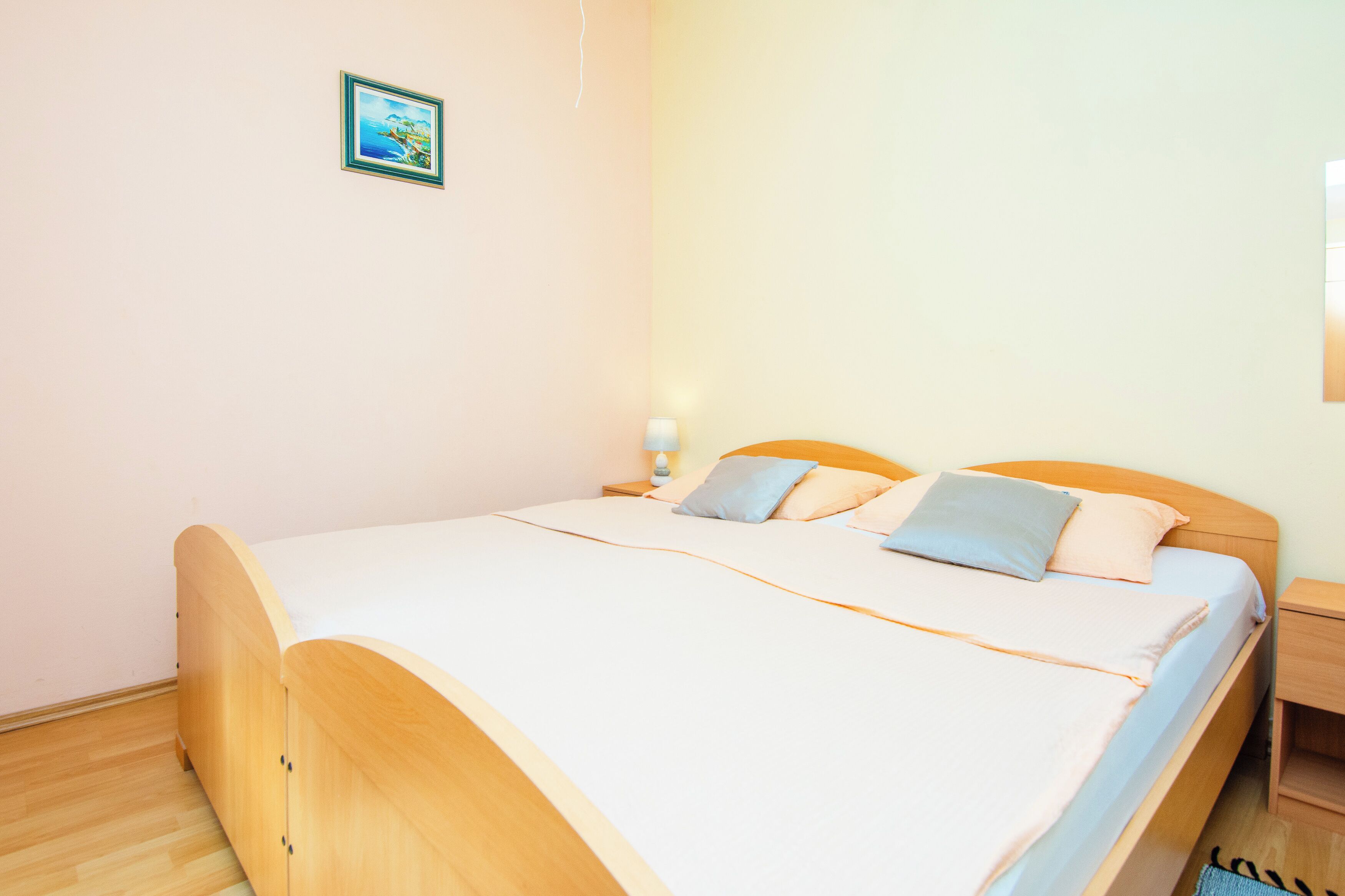 A single bed with a wooden frame, white bedding, and two pillows. There is a small bedside table with a lamp and a picture on the wall.