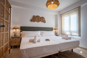 A neatly made bed with white linens and two rolled towels, a bedside table with a lamp, a wall-mounted decorative piece, and a large window with sheer curtains. - A040 PEÑON DE IFACH 6º A (Calp)