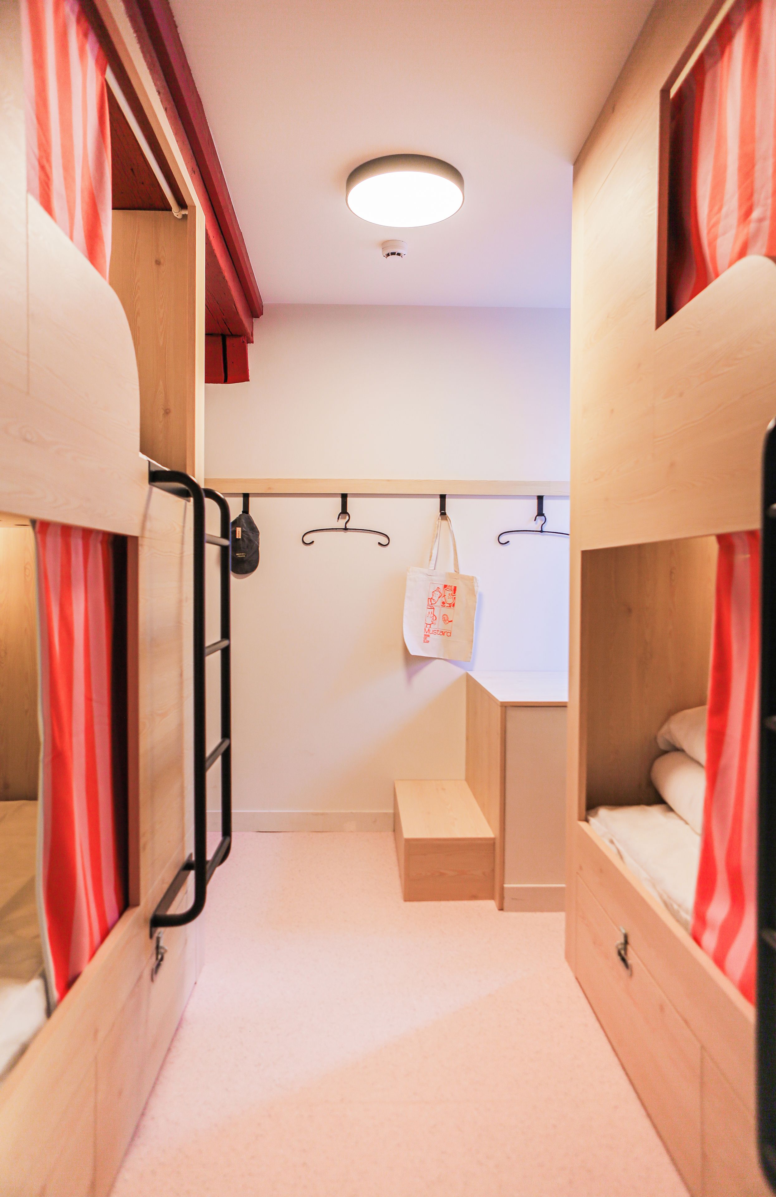 Habitación pequeña con literas, un banco, un bolso colgado y una lámpara.