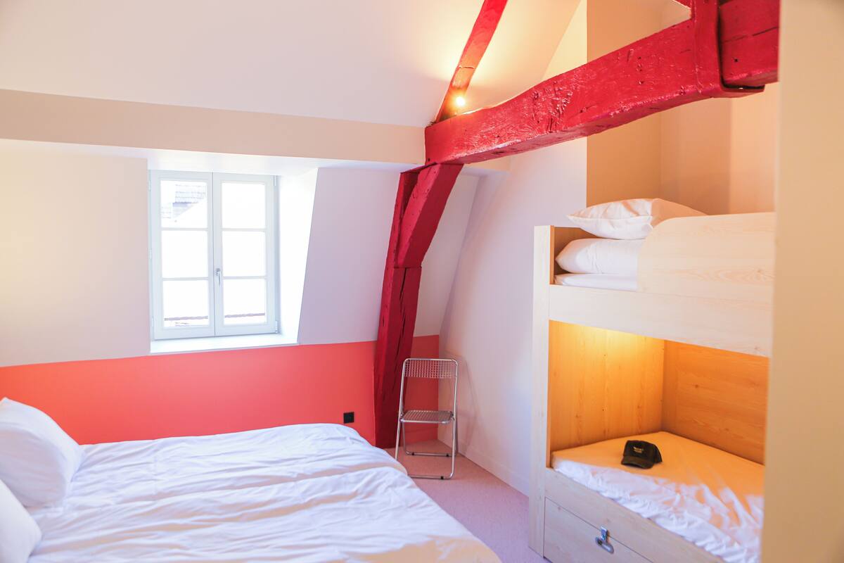 Ein Zimmer mit Etagenbett, einem Einzelbett, einem Fenster und einer roten Balkenkonstruktion.