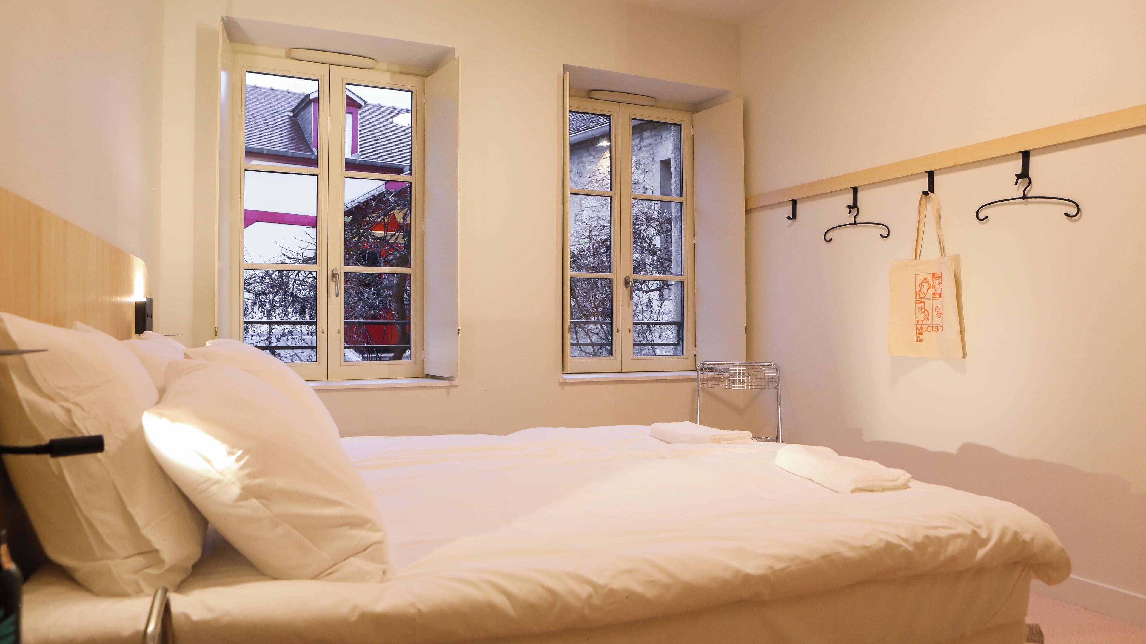Ein Hotelzimmer mit einem Bett, zwei Fenstern, einem Sessel und einem Kleiderhaken mit einer Tasche.