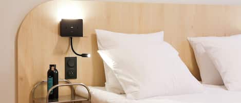 A hotel room with a wooden headboard, a bedside table with a bottle, a lamp, and a bed with white bedding.