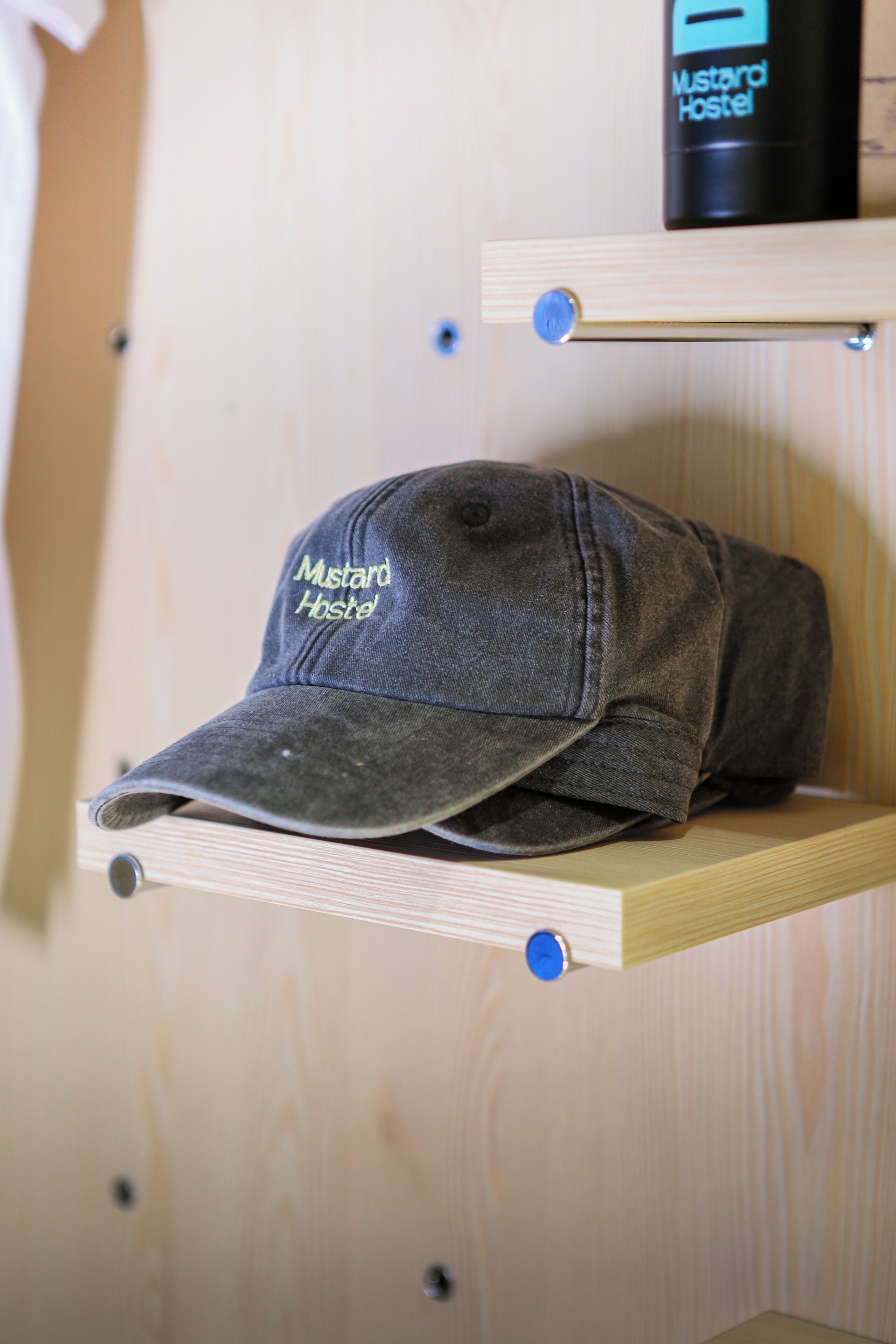 Una gorra gris con el nombre 'Mustard Hostel' bordado, sobre una estantería de madera.