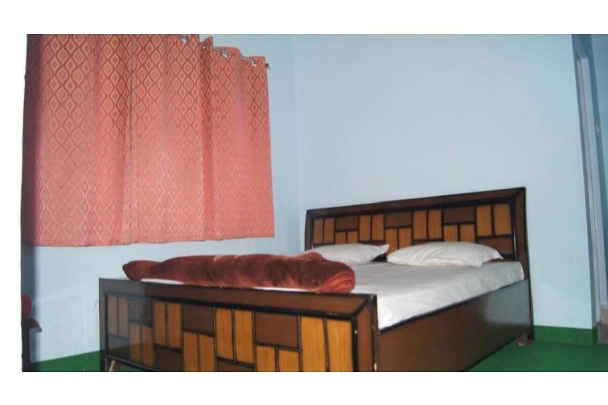 Una cama individual con estructura de madera y una colcha roja, una ventana con cortina estampada, y un suelo verde.