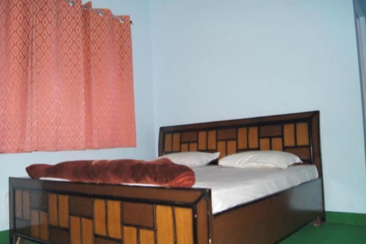 A single bed with a wooden frame and a red blanket, a window with a patterned curtain, and a green floor.