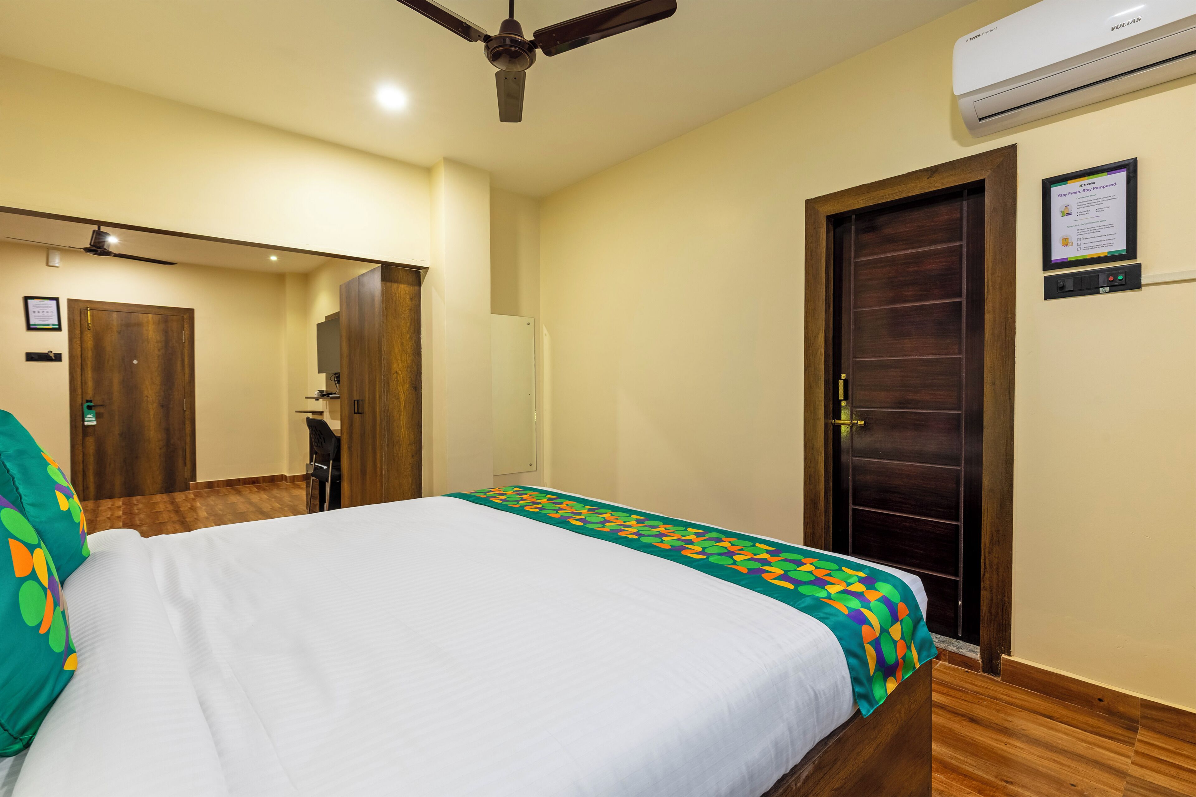 A hotel room with a bed, a ceiling fan, an air conditioning unit, a wooden door, and a mirror.