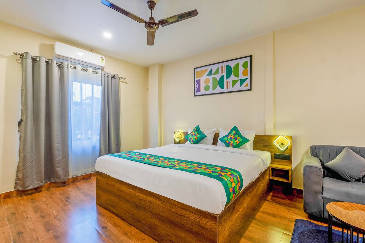 A bedroom with a bed, curtains, a ceiling fan, a wall art piece, a bedside table with a lamp, and a sofa.