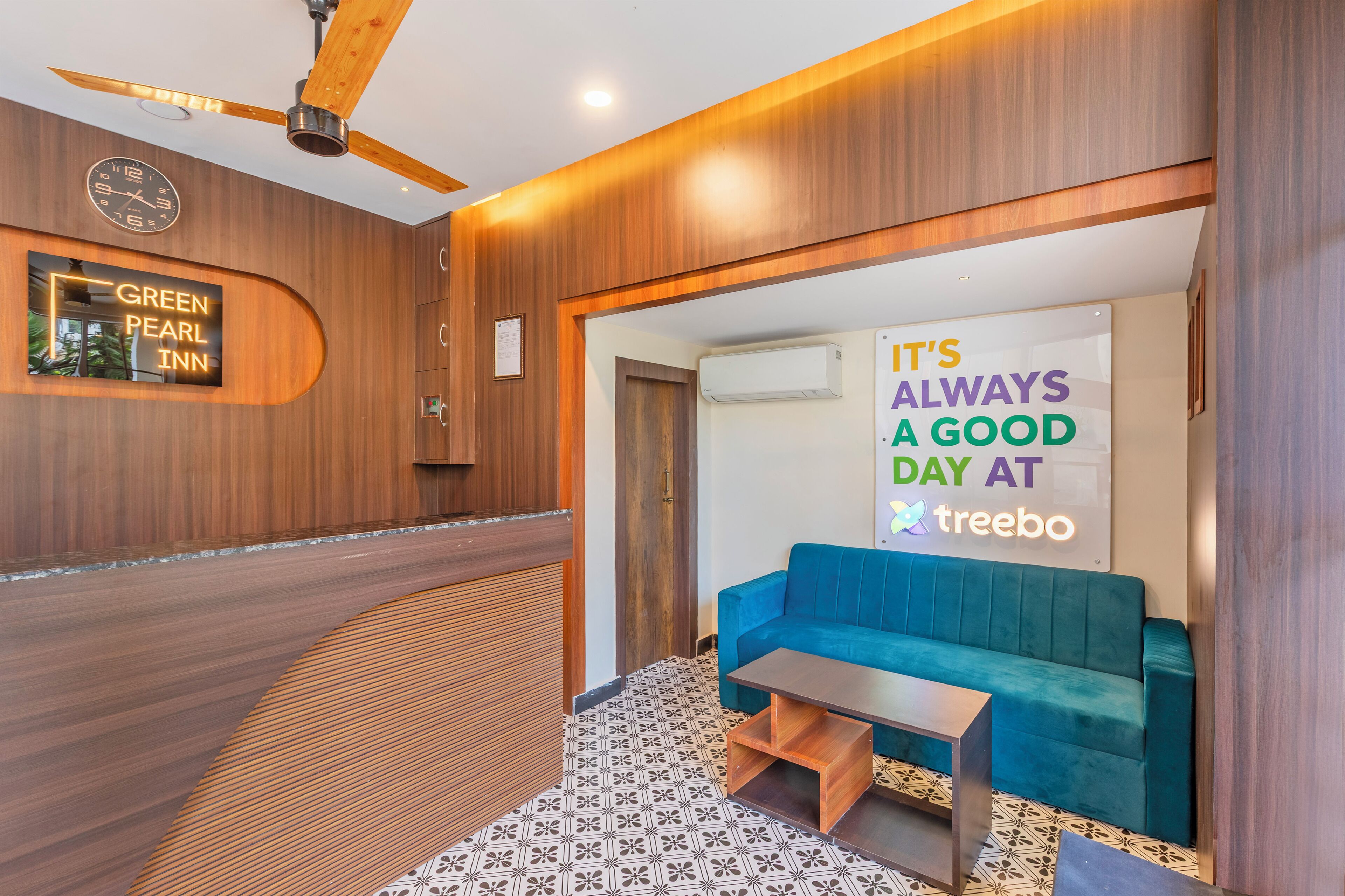A hotel lobby with a wooden reception desk, a blue sofa, a clock, and a sign that reads 'It's always a good day at treebo.'
