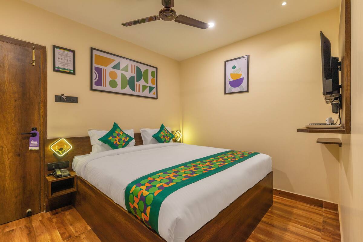 A hotel room with a bed, wooden flooring, a ceiling fan, artwork on the walls, and a television mounted on the wall.