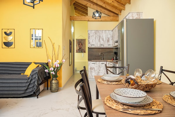 A dining area with a wooden table set for four, surrounded by chairs. A kitchen with wooden cabinets and a refrigerator is visible in the background. - Sirius House (Mitilinii)