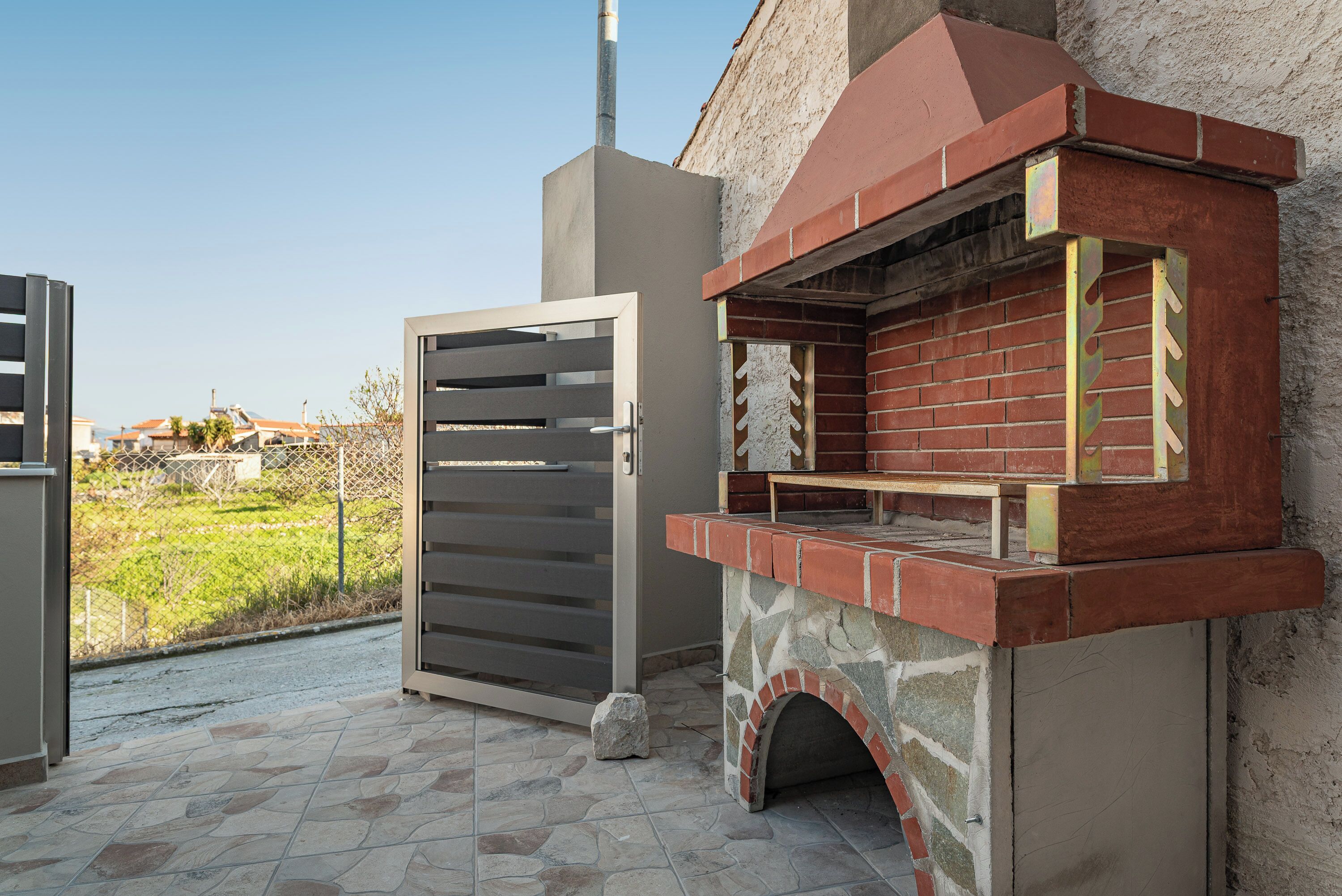 An outdoor kitchen with a stone and brick oven, a metal gate, and a view of a residential area.