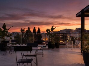 Une terrasse extérieure aménagée avec des sièges et des plantes en pot, offrant une vue sur le paysage urbain au coucher du soleil.