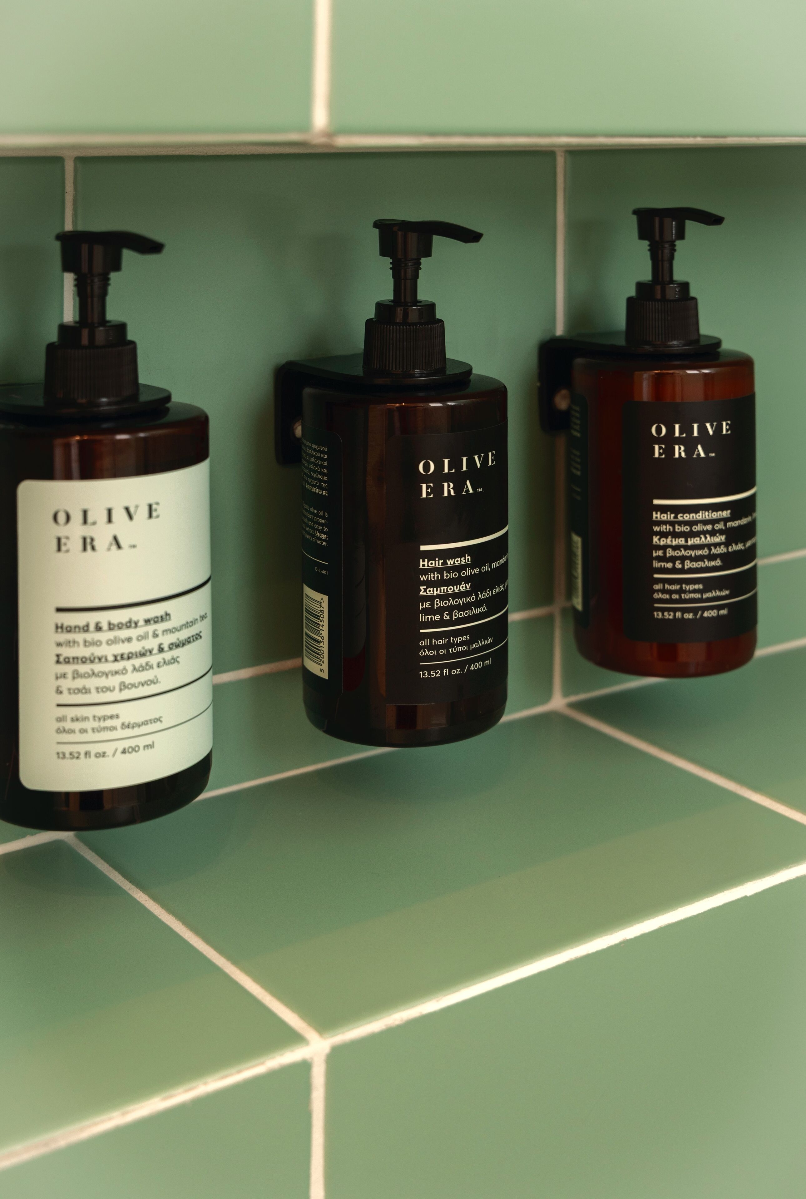 Three bottles of Olive Era hair wash and conditioner on a tiled wall.