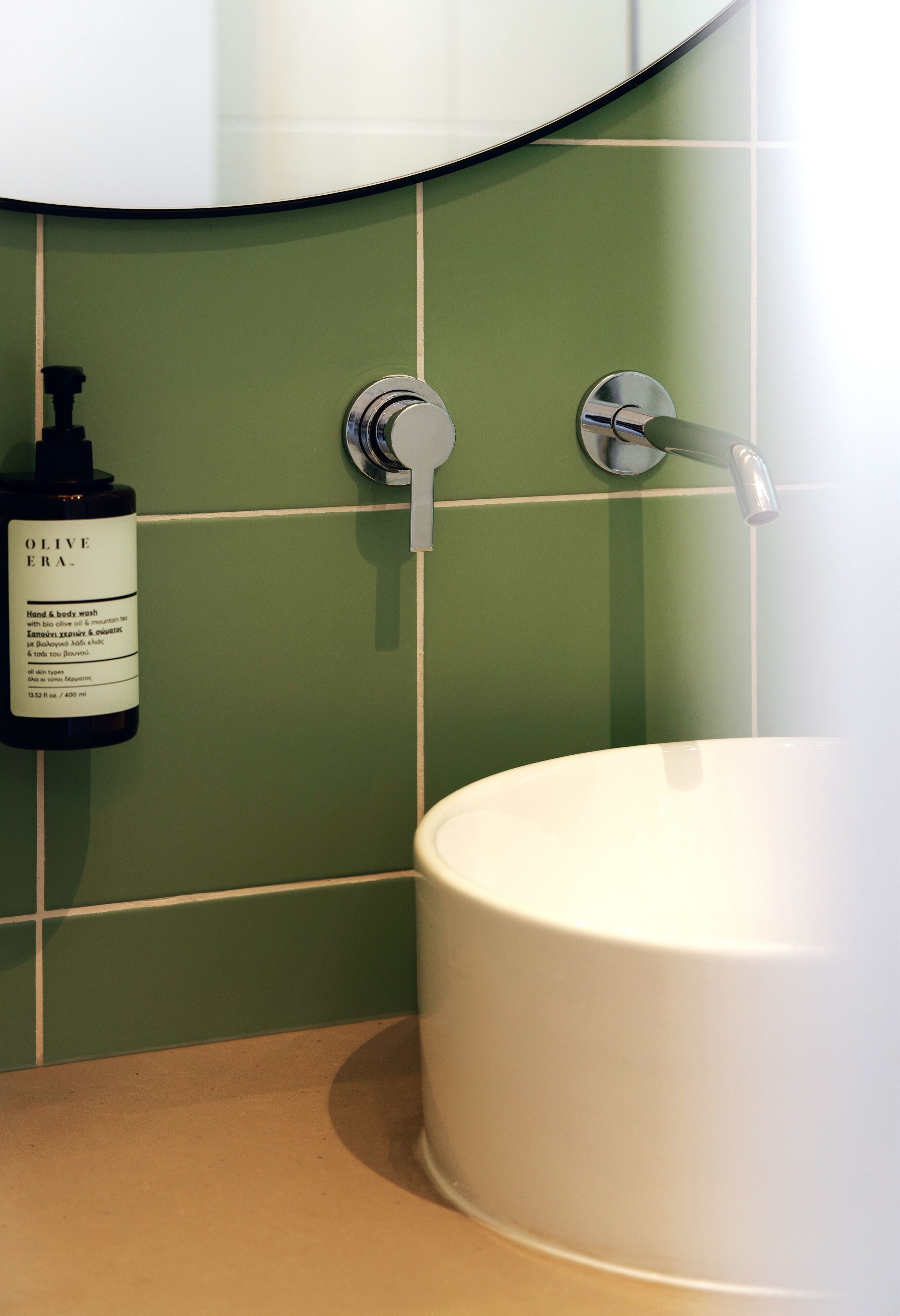 Un baño con pared de azulejos verdes, un lavamanos blanco y una botella de jabón para manos y cuerpo.