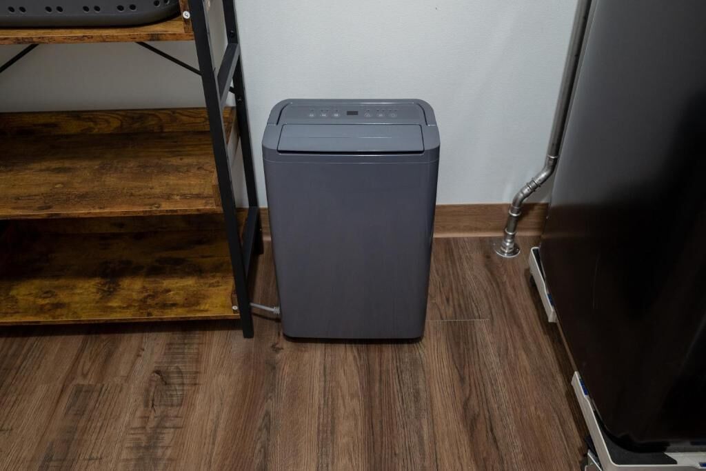 A compact, gray, wall-mounted laundry machine in a corner with a wooden floor and a metal shelf above.