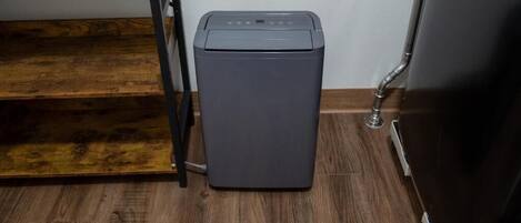A compact, gray, wall-mounted laundry machine in a corner with a wooden floor and a metal shelf above.