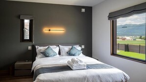 A bedroom with a large bed, a nightstand, a wall-mounted light, a window with a view of the outdoors, and a small mirror. - Furano Torinuma VILLA (Furano)