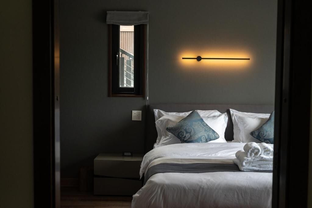 Habitación de hotel con cama, almohadas, una mesita de noche y una lámpara fijada a la pared.