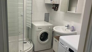 Une petite salle de bain dotée d’une douche à parois de verre, d’un lave-linge à chargement frontal, d’un lavabo avec distributeur de savon et d’un plan de travail sur lequel est posé le lave-linge.