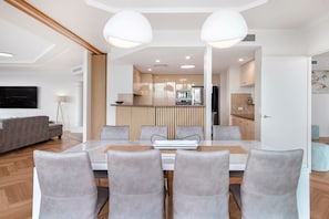 A modern living room with a dining area, a kitchen in the background, and a flat-screen TV mounted on the wall.
