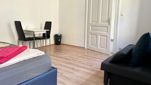 Ein Zimmer mit einem Bett, einem Schreibtisch mit zwei Stühlen, einer schwarzen Mülltonne und einer weißen Tür.