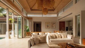 A modern living room with a sofa, a coffee table, and a dining area. There is a large window providing natural light and a view of the outdoors. - Exclusive villa in Botanica The Nature (Choeng Thale)