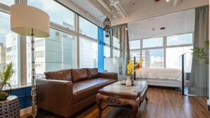 A modern room with a brown leather sofa, a wooden coffee table, and a bed with a white mattress. Large windows offer a city view. - Tung Nam Lou Art Hotel (Kowloon)