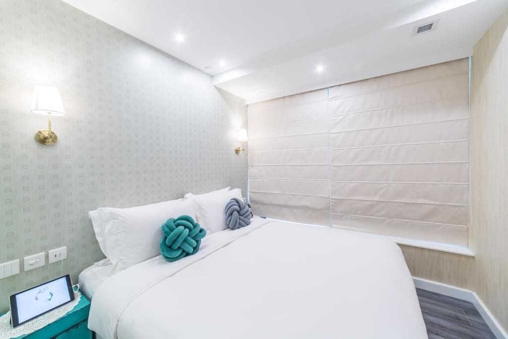 A modern hotel room with a neatly made bed, a bedside table with a lamp, and a window with blinds.