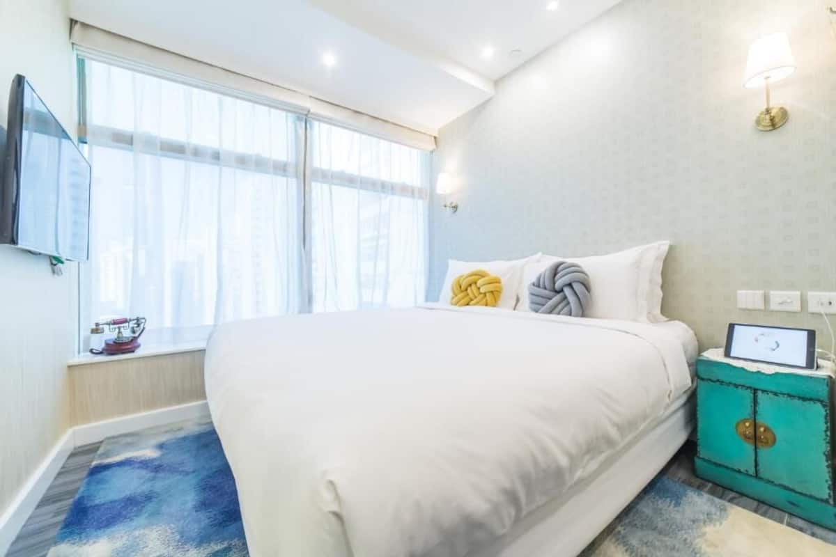 A hotel room with a large bed, a TV mounted on the wall, a window with sheer curtains, and a small table with a lamp.